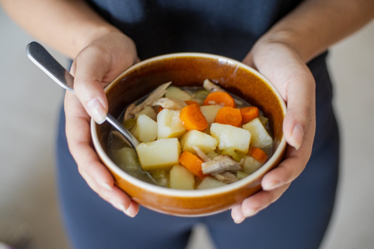 A warm bowl of homemade vegetable soup held by hands, showcasing carrots, potatoes, and chicken.